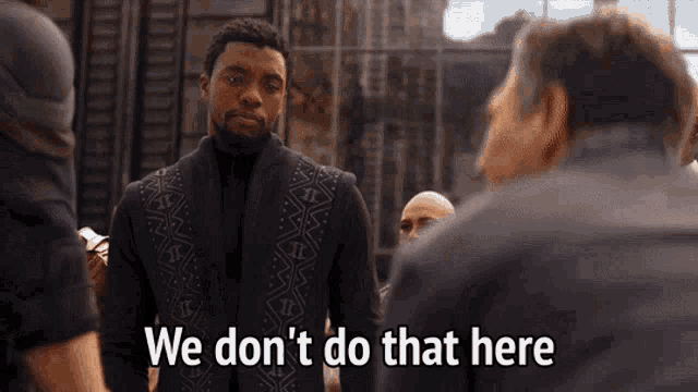 a man in a black suit is standing in front of a group of men and says we don 't do that here .