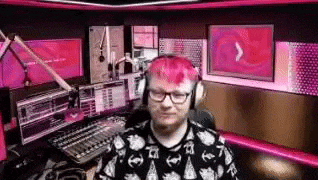 a man with pink hair and glasses is sitting in a studio wearing headphones .
