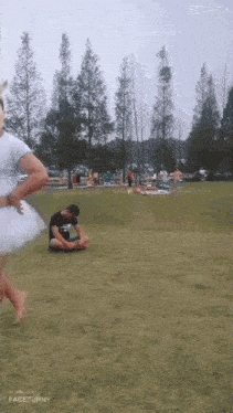 a man in a black shirt sits on the grass while a woman in a white tutu runs across the field