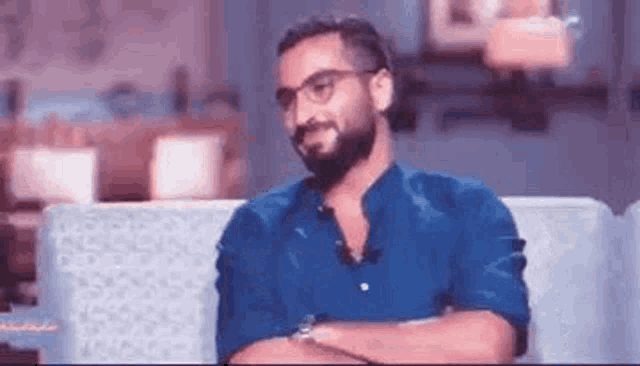 a man with a beard wearing glasses is sitting on a couch with his arms crossed .