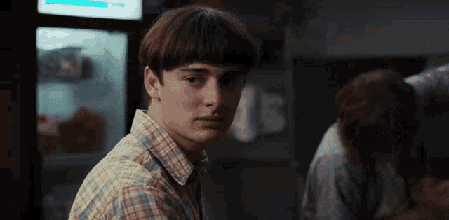a young man in a plaid shirt is looking at the camera while standing in front of a refrigerator .