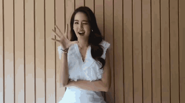 a woman in a white dress is sitting in front of a wooden wall and waving at the camera .