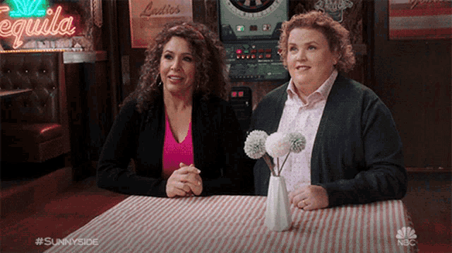 two women sitting at a table with a vase of flowers in front of a tequila sign