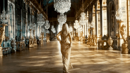 a woman in a long gold dress is walking through a hallway filled with statues and chandeliers