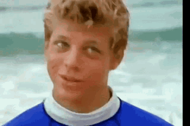 a young man is standing on the beach wearing a blue shirt and looking at the camera .
