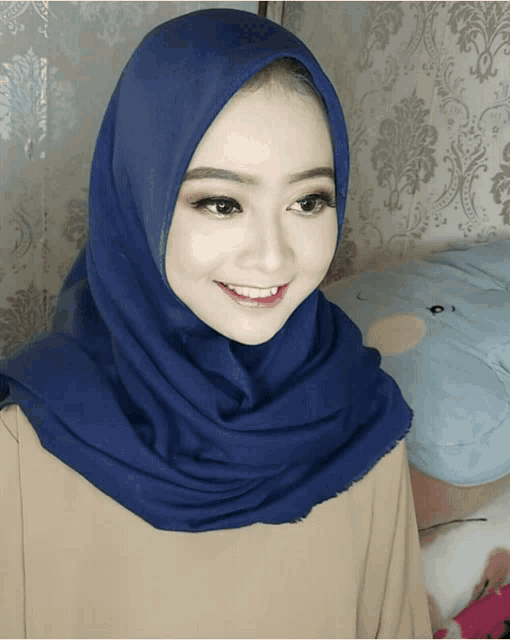 a woman wearing a blue hijab smiles in front of a wall