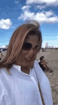 a woman wearing sunglasses and a white shirt is standing on a beach with her hair blowing in the wind .