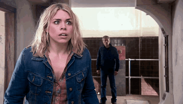 a woman in a denim jacket stands in a hallway with a man standing behind her