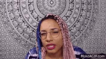 a woman with pink braids and glasses is making a funny face in front of a mandala .