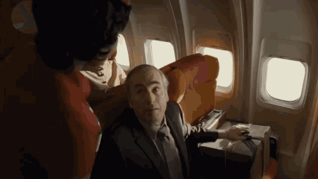 a man in a suit and tie is sitting on an airplane with a stewardess standing next to him