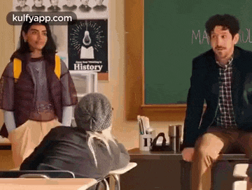 a man is sitting at a desk in front of a chalkboard in a classroom talking to a woman .