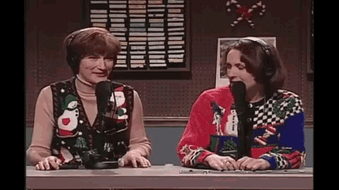 two women wearing ugly christmas sweaters are sitting at a table with microphones .
