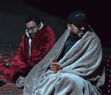 a man wearing a tommy hilfiger hat sits next to a man wrapped in a blanket