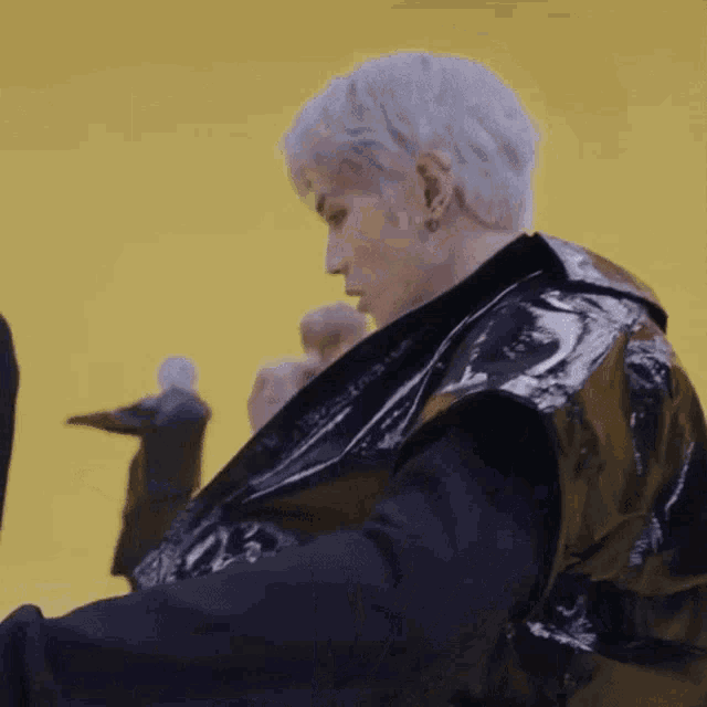 a man with white hair and a black jacket is standing in front of a yellow wall .