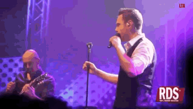 a man is singing into a microphone while another man plays a guitar on stage .