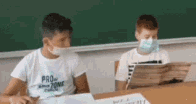 two young boys wearing face masks are sitting at a desk in a classroom .