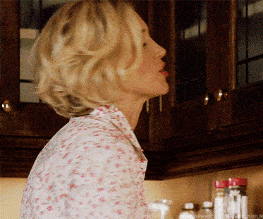 a woman in a floral shirt is standing in a kitchen looking up at something