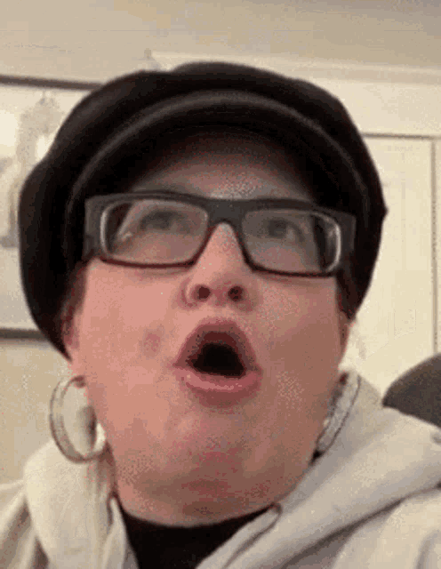 a woman wearing glasses a hat and hoop earrings is making a funny face