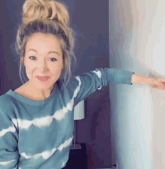 a woman in a tie dye sweater is standing next to a wall and pointing at it .