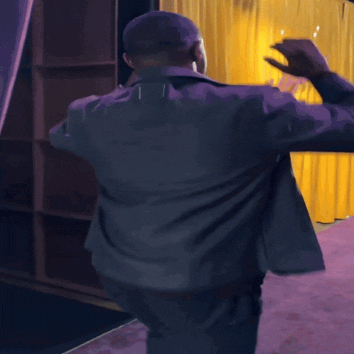 a man in a grey jacket is dancing on a purple carpet in front of a yellow curtain