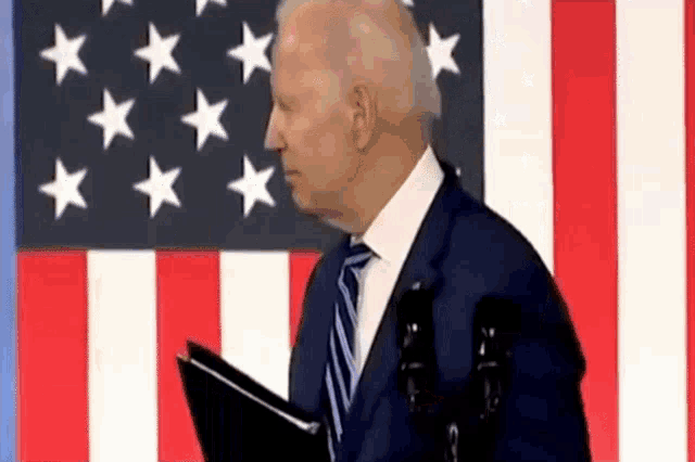 a man in a suit and tie is standing in front of an american flag and holding a folder .