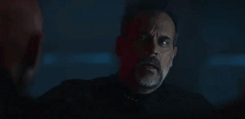 a man with a beard is sitting in a dark room talking to another man .