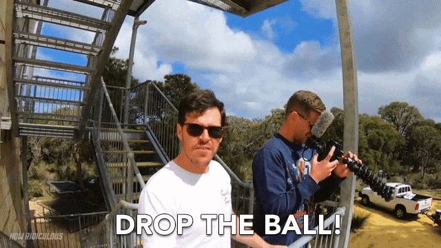 a man in sunglasses says drop the ball while another man holds a camera