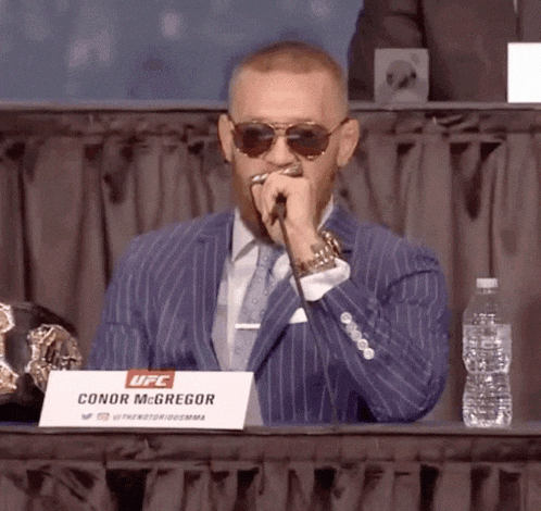 a man in a suit is sitting at a table with a sign that says conor mcgregor .