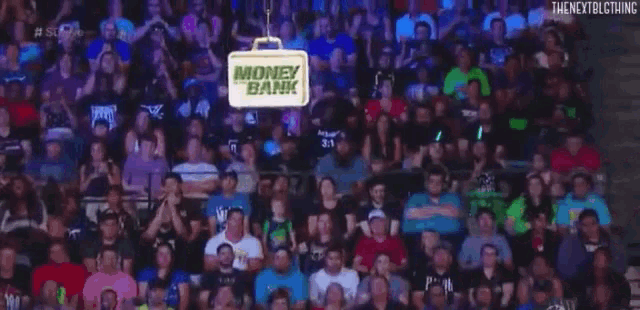 a large crowd of people are watching a wrestling match in a stadium with a money bank sign hanging from the ceiling .