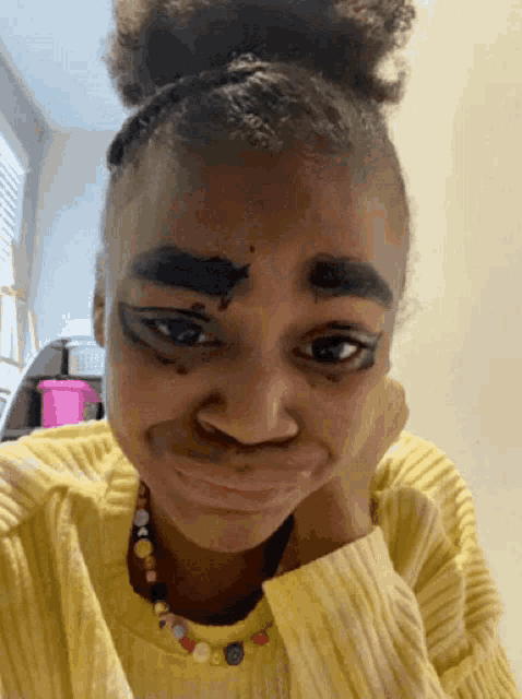 a girl with black eyebrows and a yellow sweater has her hand on her chin