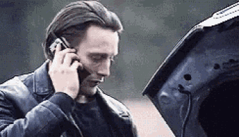 a man is talking on a cell phone while standing next to a broken down car .