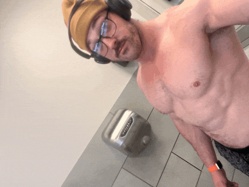 a shirtless man wearing headphones and a beanie takes a selfie in a bathroom