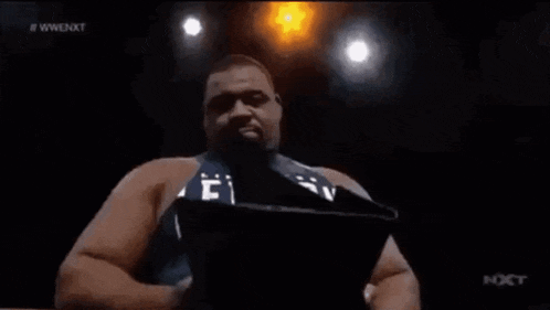 a wrestler in a blue shirt is standing in a ring with his arms in the air .