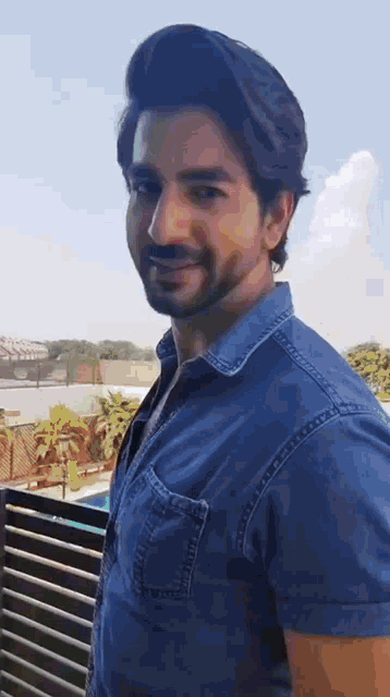 a man wearing a blue denim shirt is standing on a balcony and smiling .