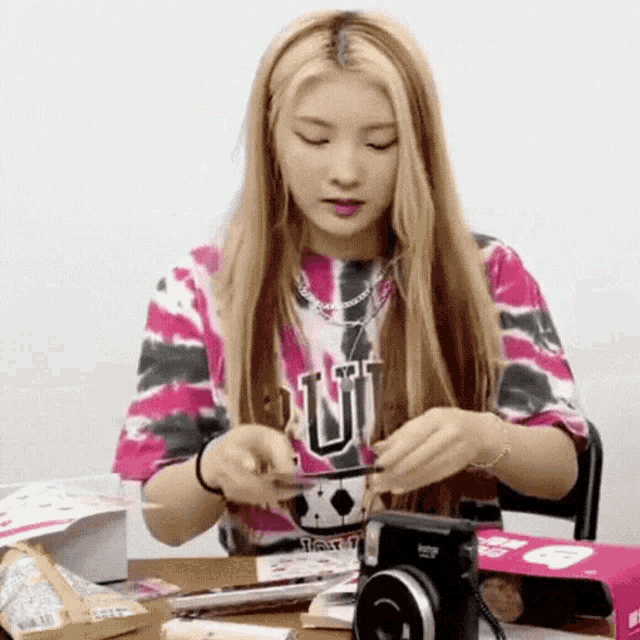 a woman wearing a tie dye shirt with the letter u on it is sitting at a table holding a camera .