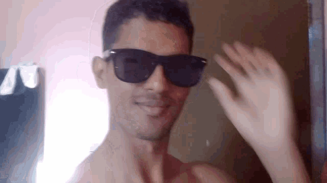a shirtless man wearing sunglasses and waving his hand