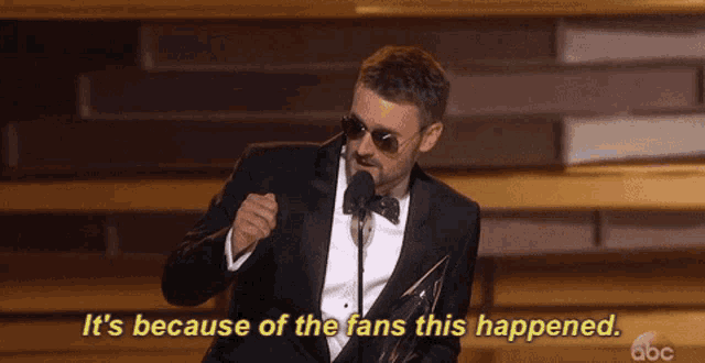 a man in a tuxedo speaking into a microphone with the words " it 's because of the fans this happened "