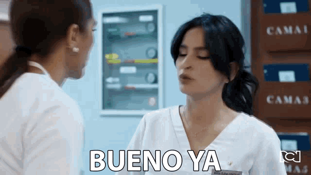 a woman in a white scrub is talking to another woman in a hospital room and says bueno ya .