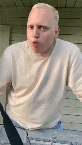 a bald man wearing a white sweater and blue jeans is standing next to a cooler and making a funny face .