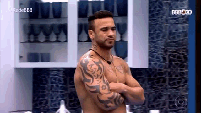a shirtless man with tattoos on his arms is standing with his arms crossed in a kitchen .