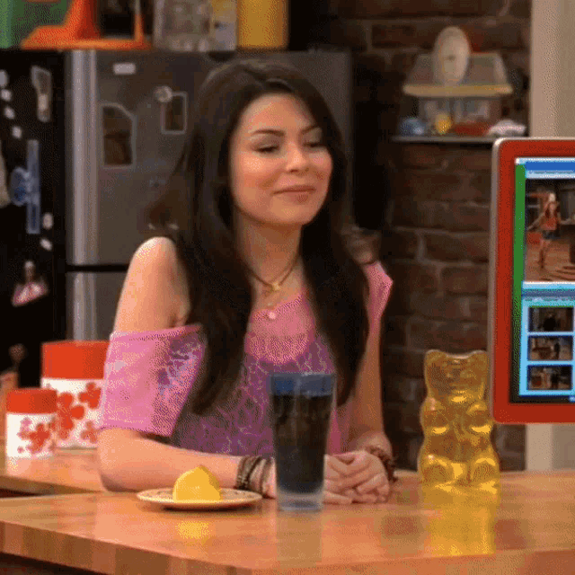 a woman in a pink top sits at a table with a gummy bear and a glass of soda