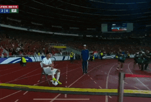 a soccer player sits on a chair in front of a scoreboard which says ksa