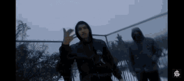 a man in a hooded jacket is standing in front of a fence .
