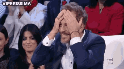 a man in a suit is covering his face with his hands while sitting in a crowd of people .