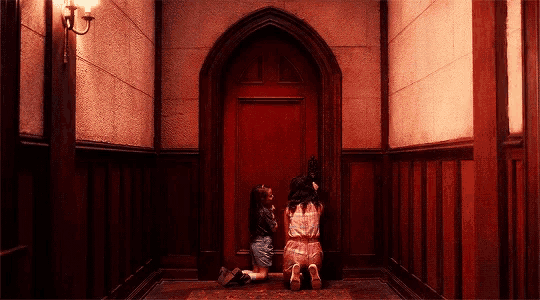 two girls are kneeling in a hallway looking at a red door .