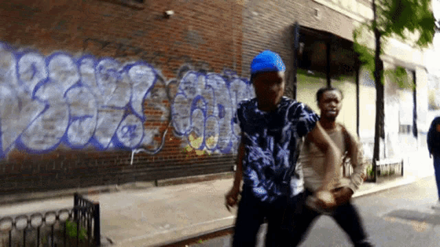 a man in a blue hat walks down a street in front of a brick wall with graffiti on it including the numbers 2236
