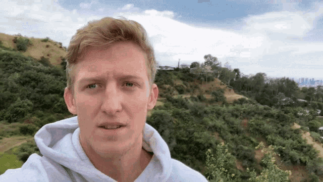 a man in a white hoodie is standing in front of a hill with trees on it