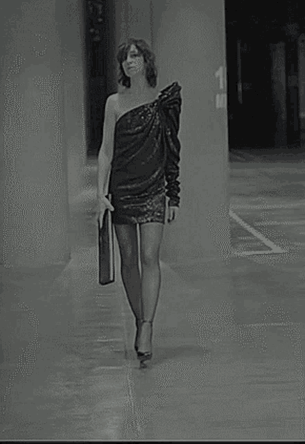 a woman in a black dress is walking down a hallway holding a black purse .