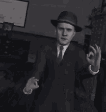 a black and white photo of a man in a suit and hat making an ok sign .