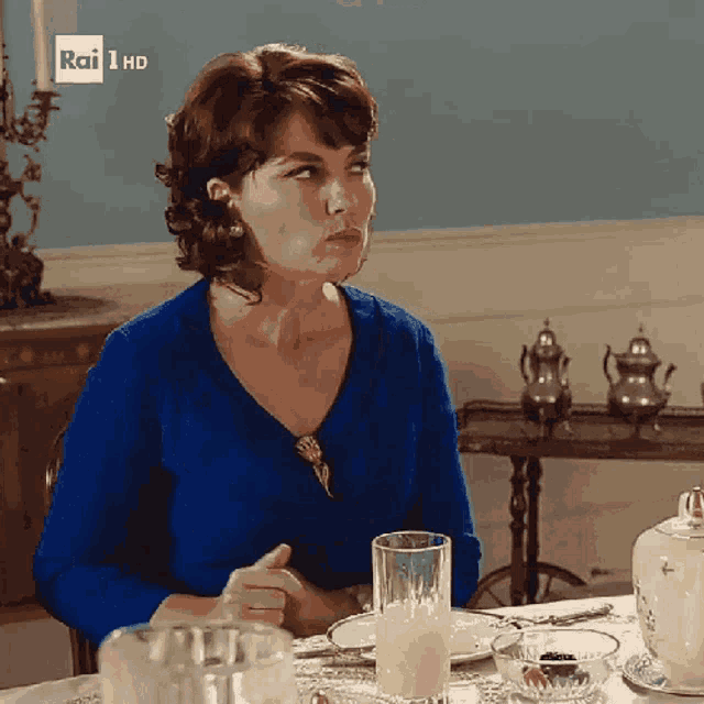 a woman in a blue sweater sits at a table with a glass of milk in front of her